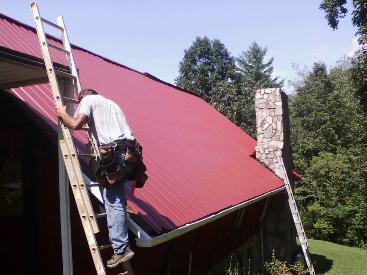 Roofer on a ladder beside a red metal roof and stone chimney — Soffit & Fascia Repair by Roofing Brothers in Kingston